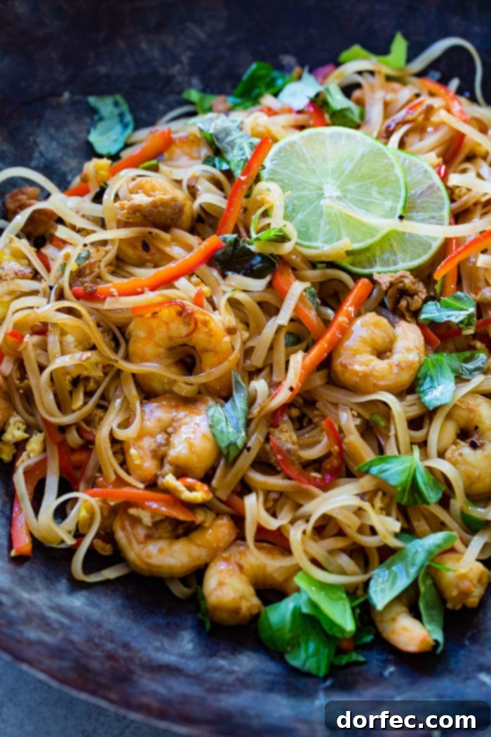 A photo of cooked shrimp, rice noodles, red bell peppers, and fresh basil leaves on a blue plate garnished with slices of lime.