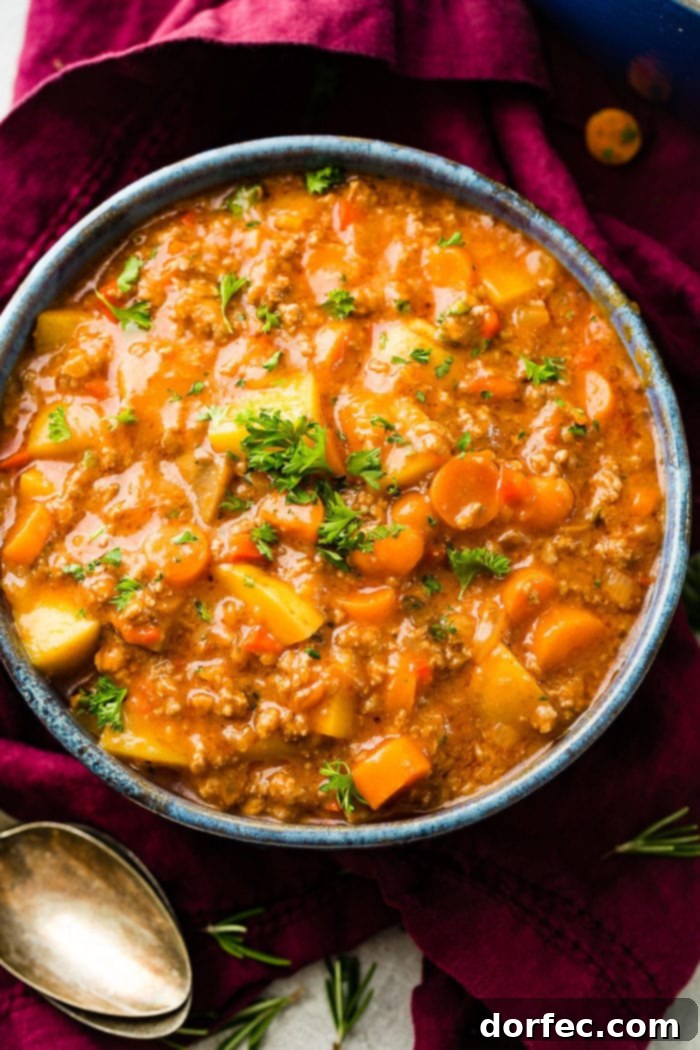 A magenta napkin with two antique spoons next to a blue-rimmed ceramic bowl full of quick and easy ground beef stew with carrots and potatoes, garnished with fresh parsley.