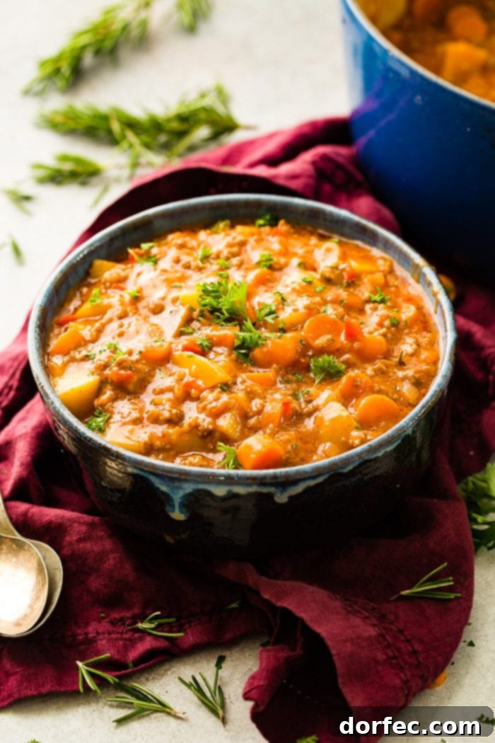 A magenta napkin with two antique spoons next to a blue-rimmed ceramic bowl full of quick and easy ground beef stew with carrots and potatoes, garnished with fresh parsley.
