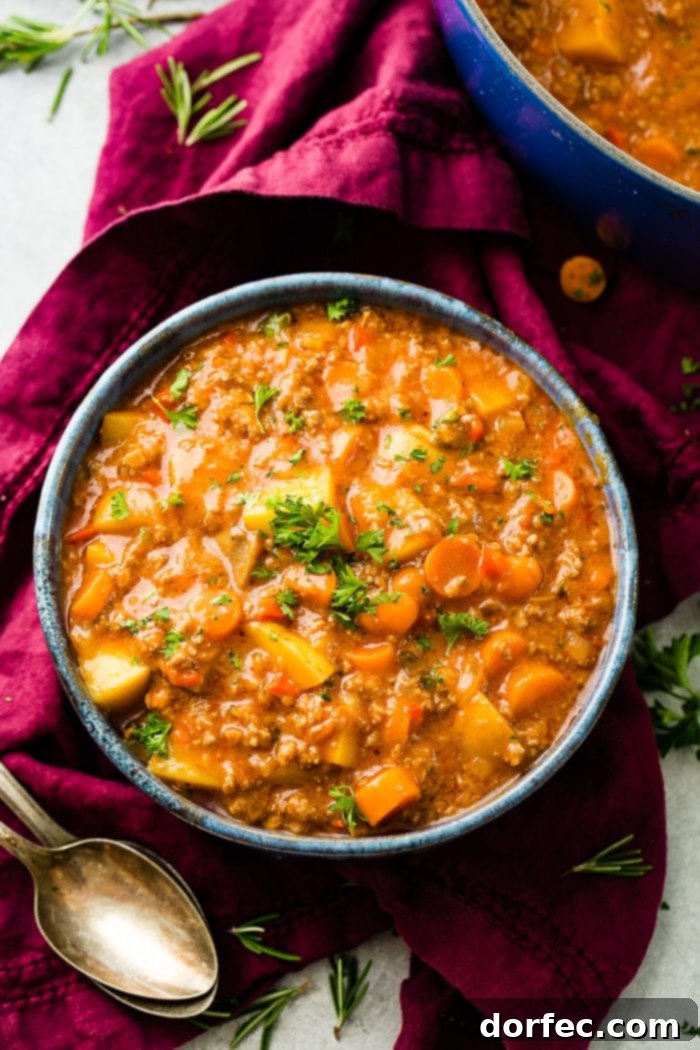 A magenta napkin with two antique spoons next to a blue-rimmed ceramic bowl full of quick and easy ground beef stew with carrots and potatoes, garnished with fresh parsley.