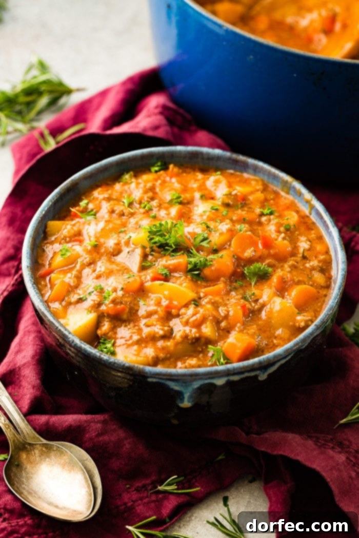 A magenta napkin with two antique spoons next to a blue-rimmed ceramic bowl full of quick and easy ground beef stew with carrots and potatoes, garnished with fresh parsley.