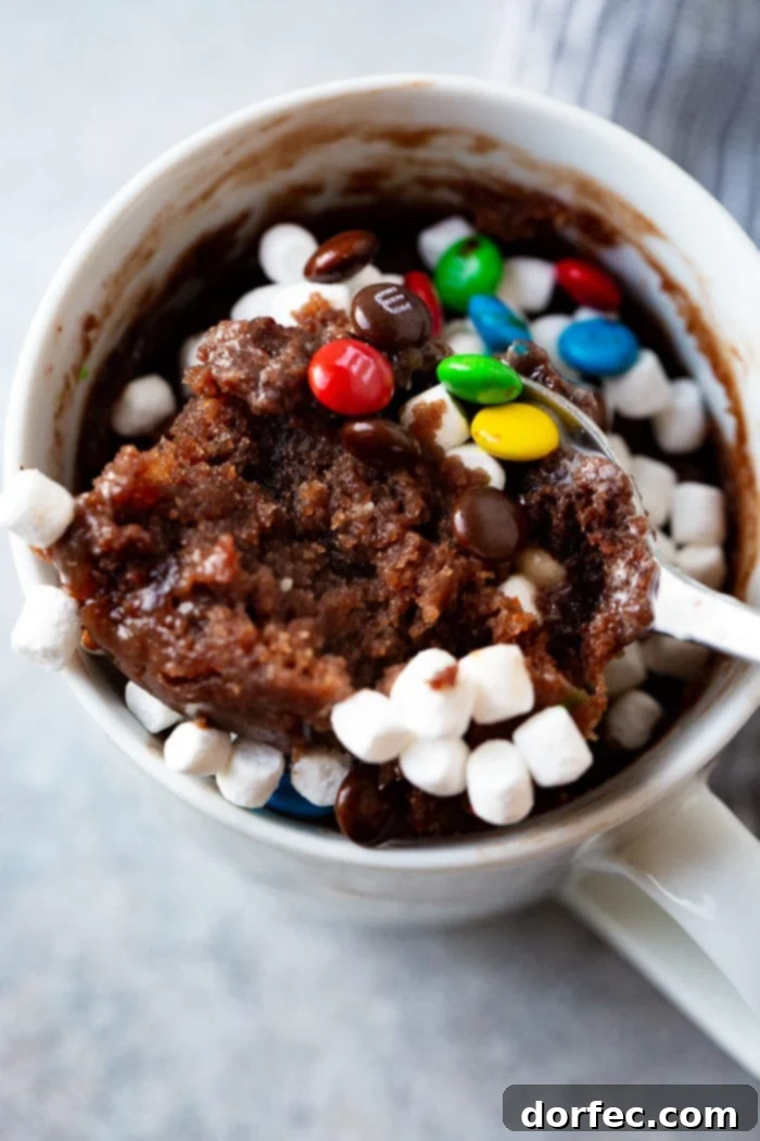 A photo of a white mug full of baked brownie topped with mini marshmallows and mini M&Ms with a spoonful being taken out