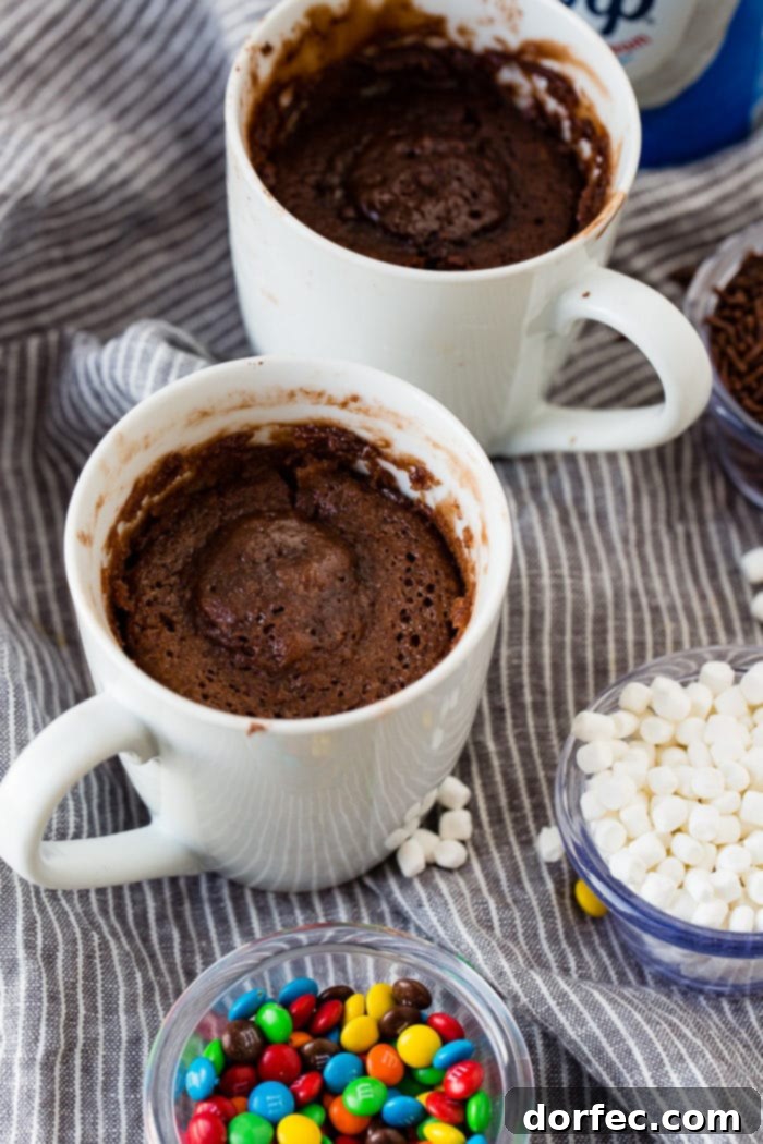 A photo of two white mugs full of cooked brownies surrounded by little bowls of sprinkles, marshmallows, and mini M&Ms