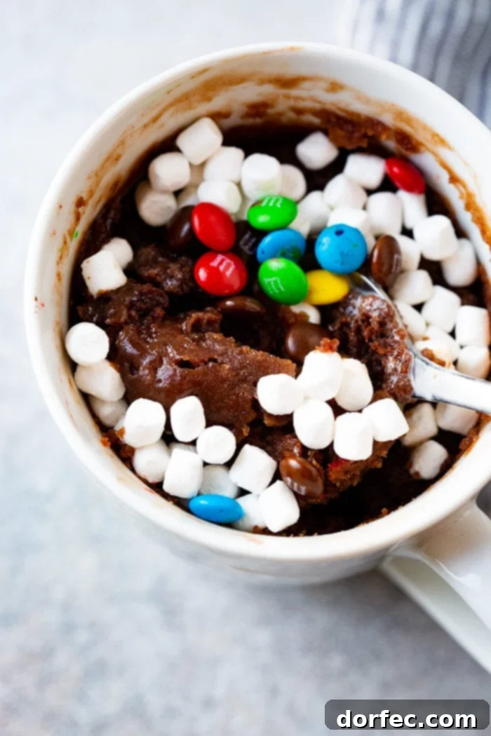 A photo of a white mug full of baked brownie topped with mini marshmallows and mini M&Ms with a spoonful being taken out