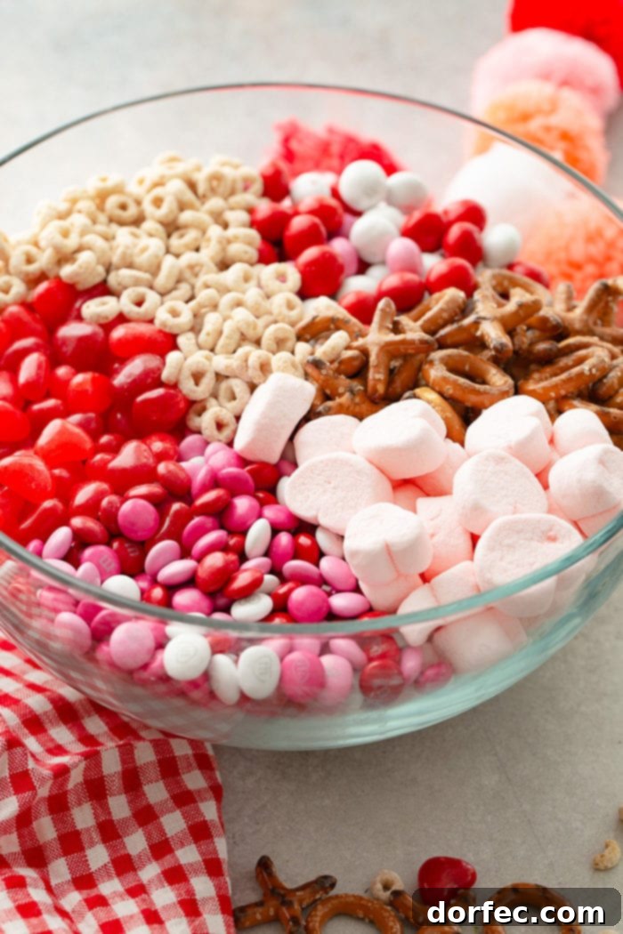 A close-up shot of the colorful ingredients for the Valentine's Snack Mix, showcasing frosted Cheerios, various M&M's, 'XO' shaped pretzels, and heart-shaped marshmallows.