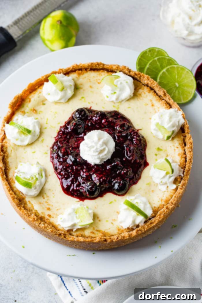 An exquisitely crafted whole lime cheesecake on a pristine white serving platter, adorned with delicate dollops of whipped cream around its edges, each crowned with a vibrant slice of fresh lime. A generous pile of rich triple berry sauce cascades over the center, featuring an additional dollop of whipped cream on top, ready to be savored.