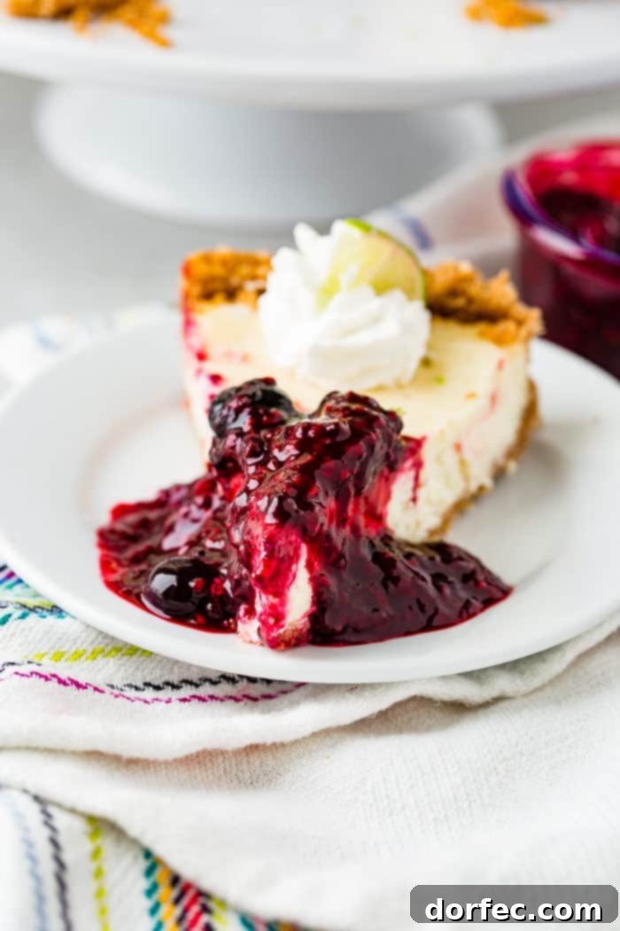 A perfect slice of lime cheesecake, sumptuously covered in triple berry sauce, sits on a white plate. A fluffy dollop of whipped cream and a delicate slice of lime complete this inviting dessert presentation.