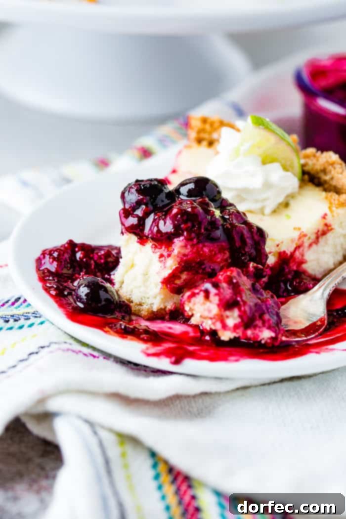 A mouth-watering close-up of a slice of lime cheesecake on a white plate, with a bite taken out, showcasing the vibrant triple berry sauce liberally dripping around it. The rich textures and colors highlight an irresistible dessert.