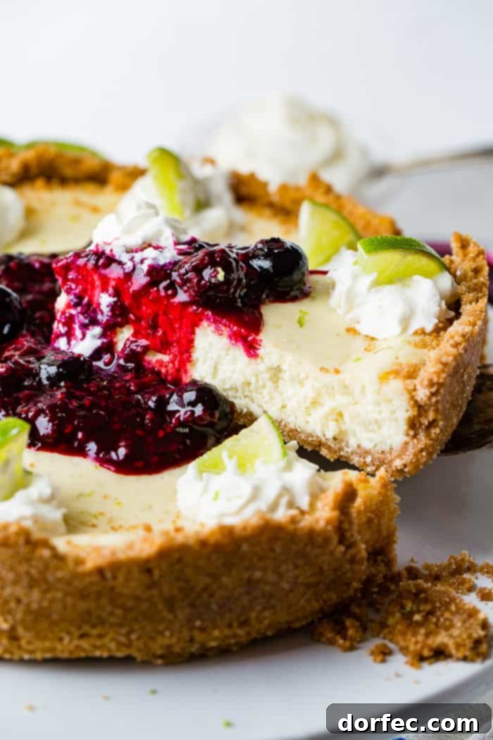A delectable slice of lime cheesecake being carefully lifted from the whole dessert, showcasing a rich triple berry sauce generously dripping down its sides. A delicate dollop of whipped cream, crowned with a vibrant slice of fresh lime, adorns the top of the slice, inviting a taste.