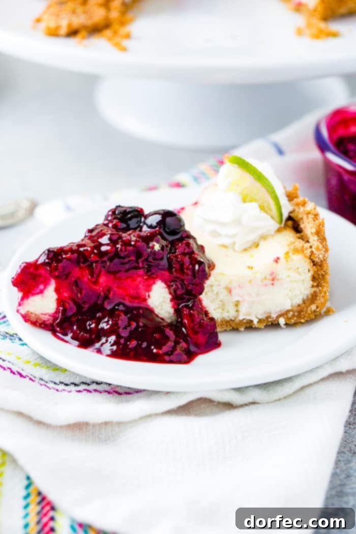 A pristine slice of lime cheesecake, generously blanketed in a vibrant triple berry sauce, rests on a white plate. It's elegantly garnished with a delicate dollop of whipped cream and a fresh lime slice, completing a picture of dessert perfection.