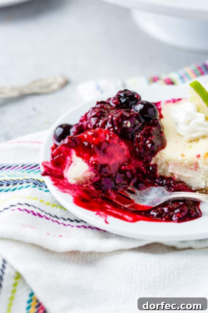 A fork delicately takes a bite from a slice of lime cheesecake on a white plate, revealing a luscious triple berry sauce cascading around it. The rich, vibrant colors and textures invite a delightful taste experience.