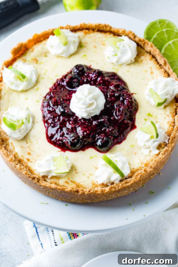 An overhead view of a magnificent whole lime cheesecake, elegantly presented on a white serving platter. Adorning its edges are delicate dollops of whipped cream, each graced with a tiny, vibrant slice of fresh lime. The center is a luscious mound of triple berry sauce, topped with another perfect dollop of whipped cream, creating a feast for the eyes and palate.
