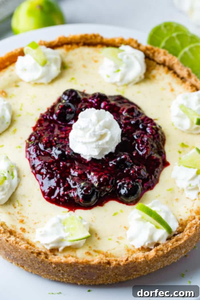 A detailed, overhead shot of a luscious whole lime cheesecake on a white serving platter. It's elegantly decorated with uniform dollops of whipped cream around the perimeter, each holding a bright green slice of lime. A vibrant, generous pile of triple berry sauce crowns the center, topped with a final perfect dollop of whipped cream, creating an appetizing and beautiful dessert.