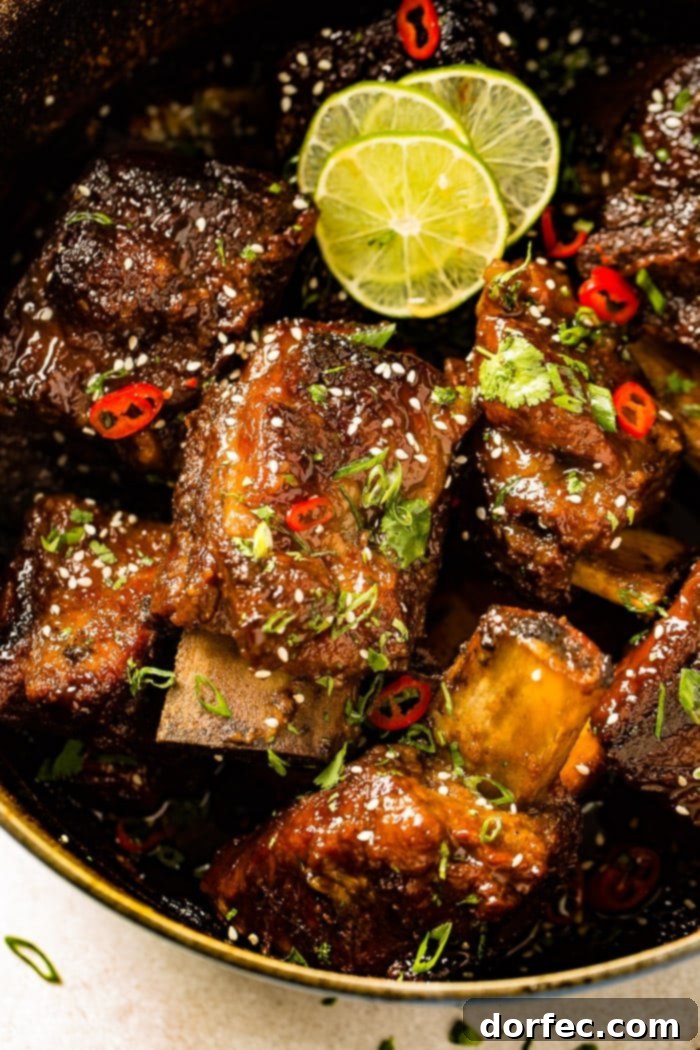 Fall-Off-The-Bone Oven Braised Korean Beef Short Ribs 3 A photo of several braised short ribs still in the dutch oven garnished with sesame seeds, red chile peppers, cilantro, green onions, and lime slices.