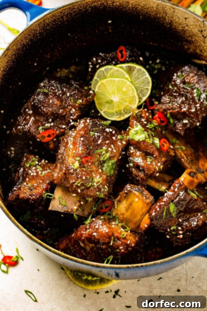 Fall-Off-The-Bone Oven Braised Korean Beef Short Ribs 7 A photo of several braised short ribs still in the dutch oven garnished with sesame seeds, red chile peppers, cilantro, green onions, and lime slices.