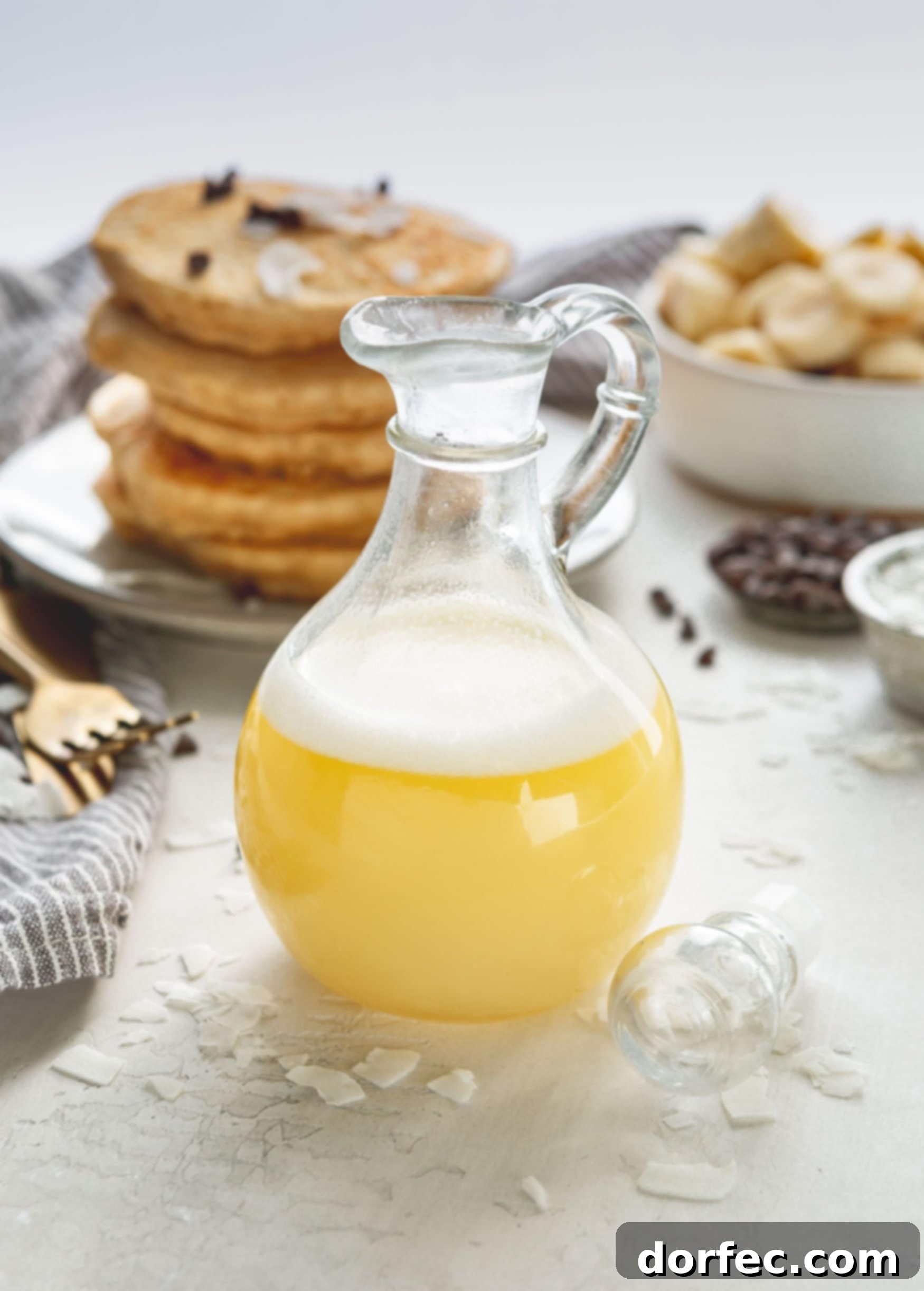Tropical Coconut Buttermilk Syrup 2 A syrup container with coconut buttermilk syrup. The syrup is light yellow in color and has a bit of white foam on top. There is a stack of pancakes in the background and a few chocolate chips are scattered on top.