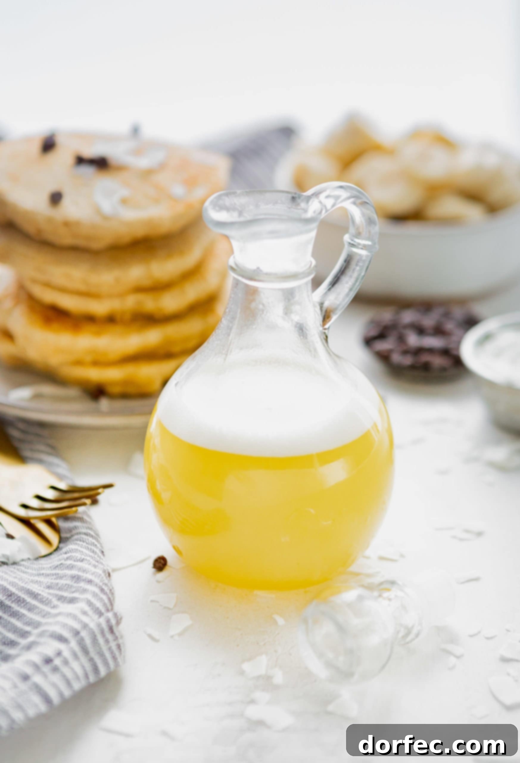 Tropical Coconut Buttermilk Syrup 3 A small syrup pitcher with coconut buttermilk syrup. The syrup is light yellow in color and has a bit of white foam on top. A stack of pancakes with chocolate chips on top and a bowl of sliced bananas are in the background.
