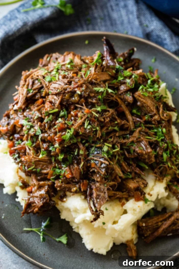 Fork-Tender Beef Roast 2 A photo of a dark gray plate with a pile of mashed potatoes on it covered in juicy shredded braised beef topped with chopped fresh parsley.