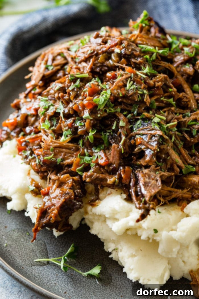Fork-Tender Beef Roast 3 A close up photo of a dark gray plate with a pile of mashed potatoes on it covered in juicy shredded braised beef topped with chopped fresh parsley.