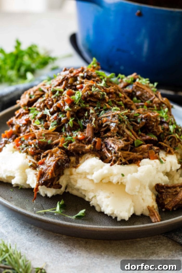 Fork-Tender Beef Roast 4 A photo of a dark gray plate with a pile of mashed potatoes on it covered in juicy shredded braised beef topped with chopped fresh parsley.