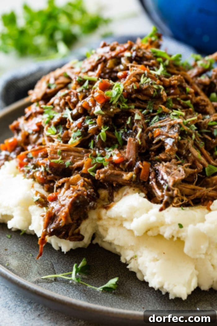 Fork-Tender Beef Roast 7 A photo of a dark gray plate with a pile of mashed potatoes on it covered in juicy shredded braised beef topped with chopped fresh parsley.