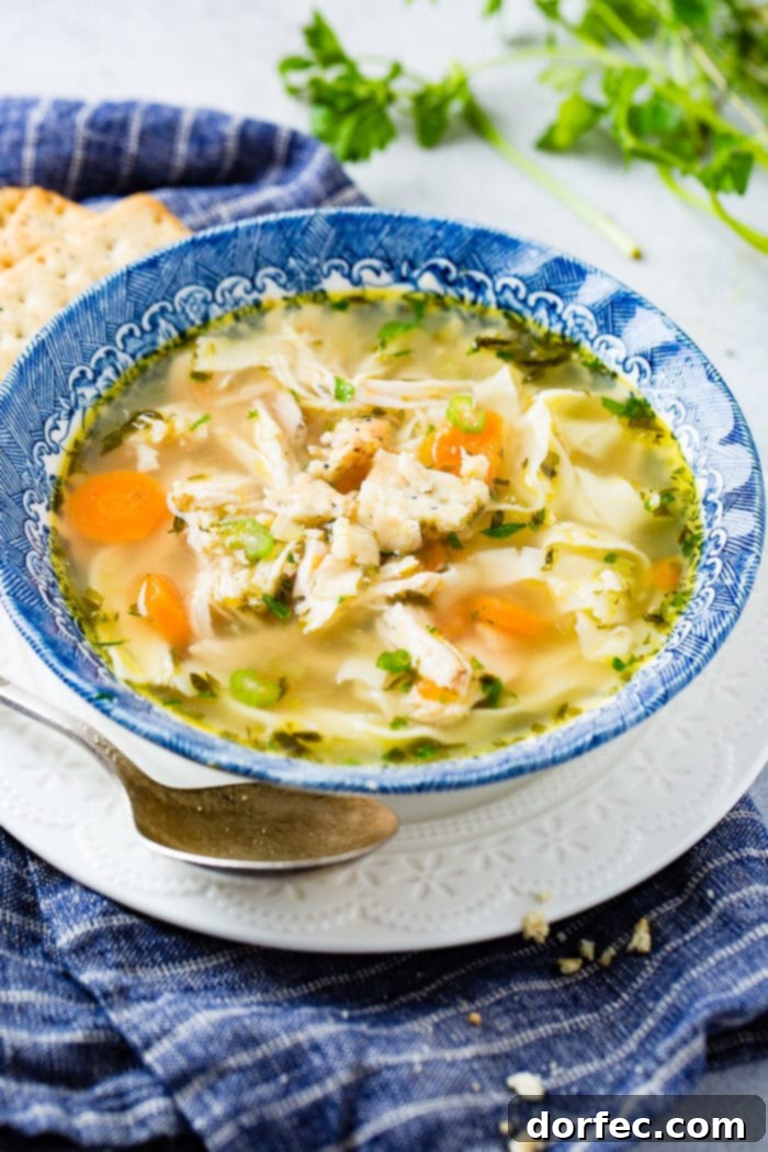 A photo of a bowl of homemade chicken noodle soup in a blue bowl sitting on a white plate with a spoon sitting by the side of the bowl.