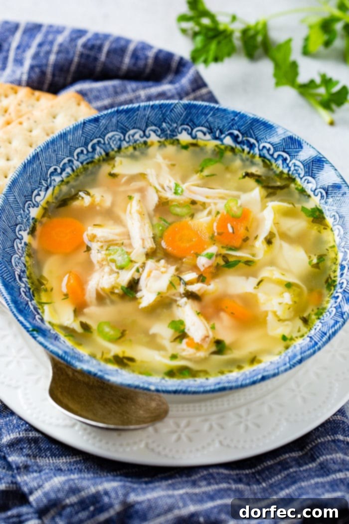A photo of a bowl of pressure cooker chicken noodle soup in a blue bowl sitting on a white plate with a spoon sitting by the side of the bowl.