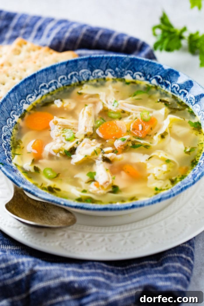 A photo of a bowl of instant pot chicken noodle soup in a blue bowl sitting on a white plate with a spoon sitting by the side of the bowl.