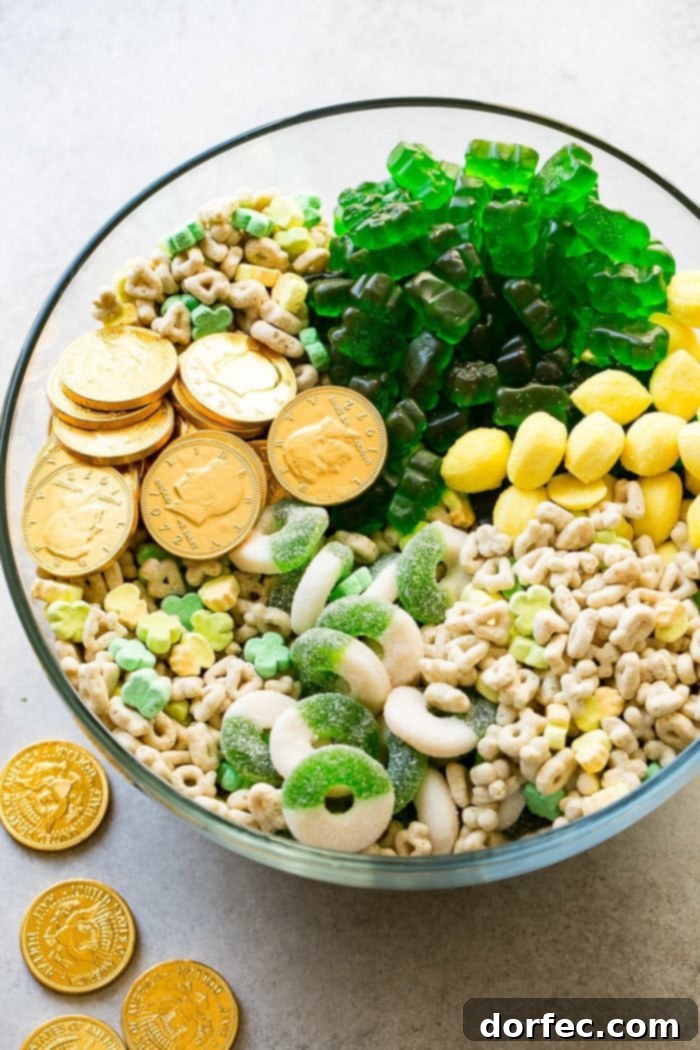 Instant Pot o' Gold Snack Mix 2 A large, clear glass bowl brimming with a colorful assortment of St. Patrick's Day themed treats. The mix includes shining chocolate gold coins, whimsical Lucky Charms cereal with its vibrant marshmallows, tangy lemon drops, chewy green apple gummy rings, and playful green gummy bears, all perfectly blended for a festive snack.