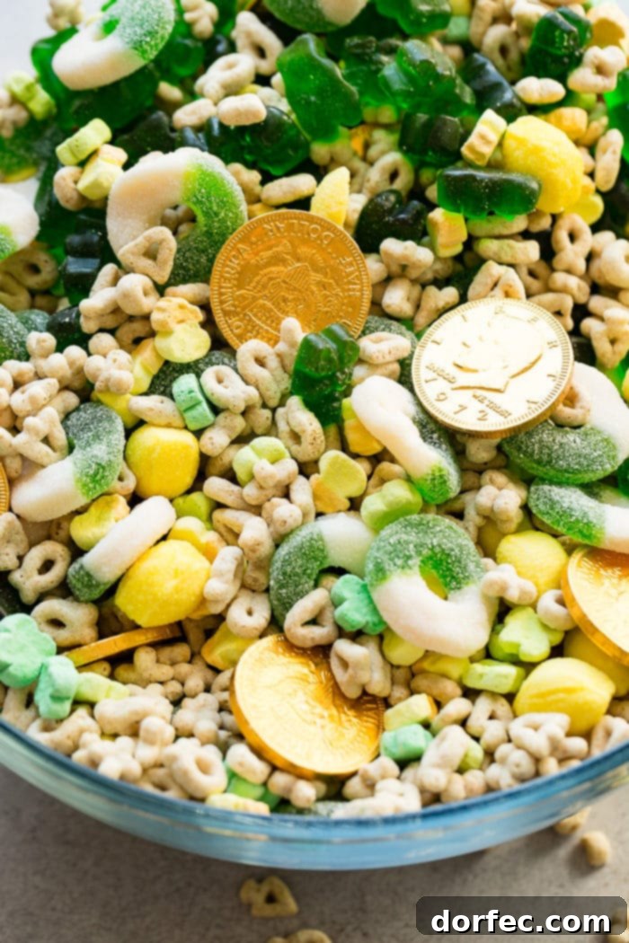 Instant Pot o' Gold Snack Mix 3 A close-up shot of a magnificent glass bowl overflowing with a visually striking St. Patrick's Day snack mix. The image prominently features a treasure trove of glistening chocolate gold coins nestled among bright green gummy candies, an abundance of colorful Lucky Charms marshmallows, and tangy lemon drops, creating an irresistible display of festive treats.