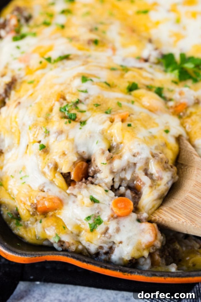 A photo of a scoop of cheesy ground beef and rice casserole being removed from an orange casts iron skillet with a wooden spoon.
