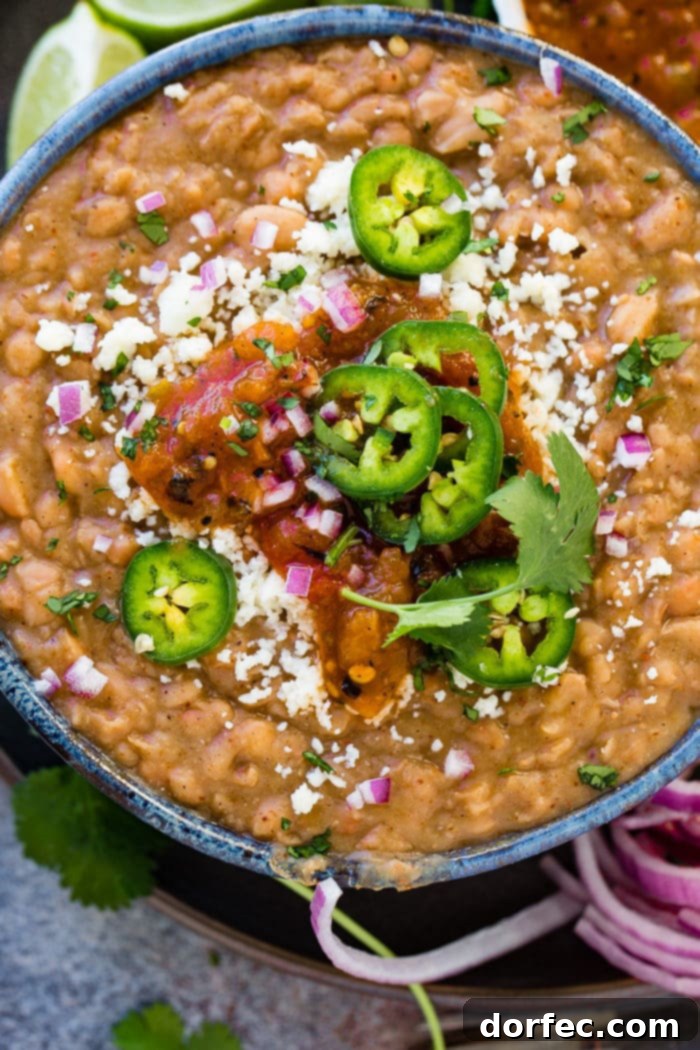 A photo of a dark blue bowl full of instant pot refried beans topped with fresh sliced jalapeno, diced purple onion, cotija cheese, and salsa.