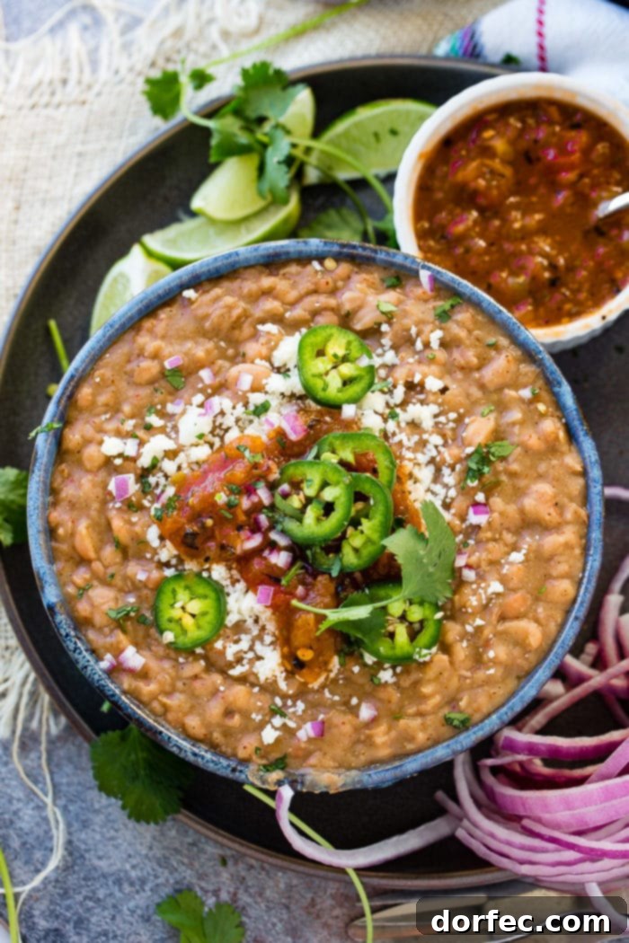 A photo of a dark blue bowl full of homemade refried beans topped with fresh sliced jalapeno, diced purple onion, cotija cheese, and and salsa. There is a cup of salsa sitting next to the bowl.