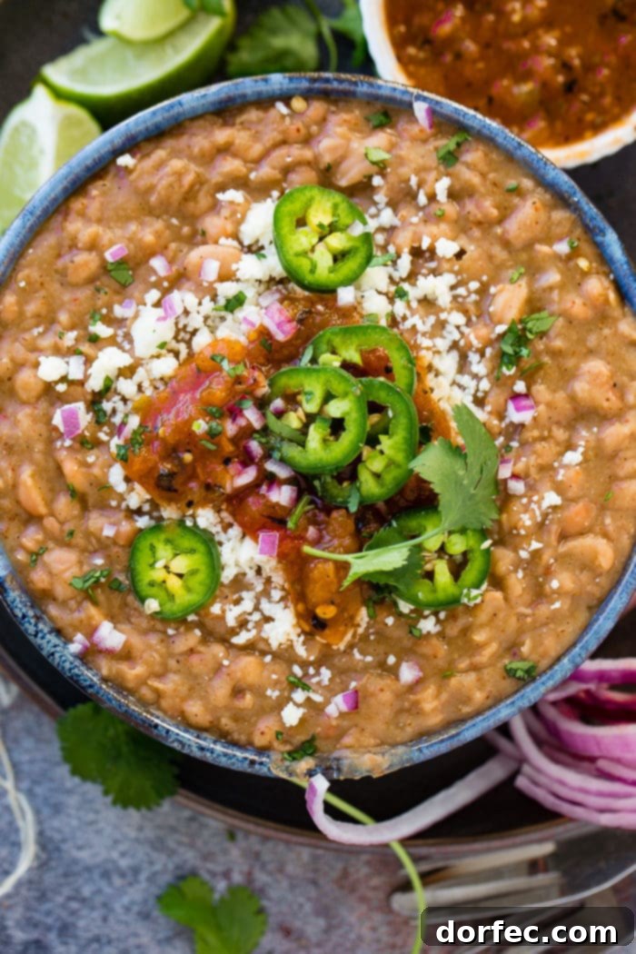 A photo of a dark blue bowl full of restaurant style refried beans topped with fresh sliced jalapeno, diced purple onion, cotija cheese, and salsa.