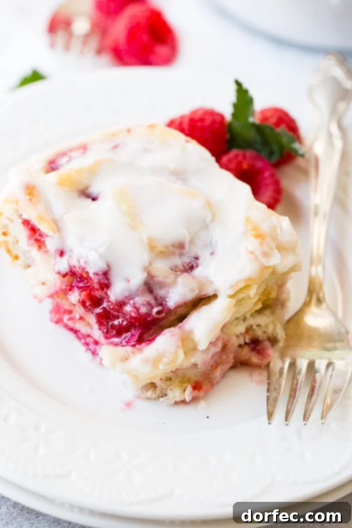 A frosted easy raspberry sweet roll on a white plate with a fork laying beside it and fresh raspberries next to it.