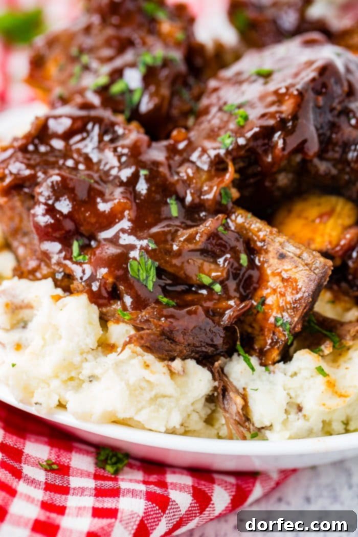 A photo of sticky bbq beef short ribs piled on top of mashed potatoes on a white plate on top of a red and white checkered cloth napkin.