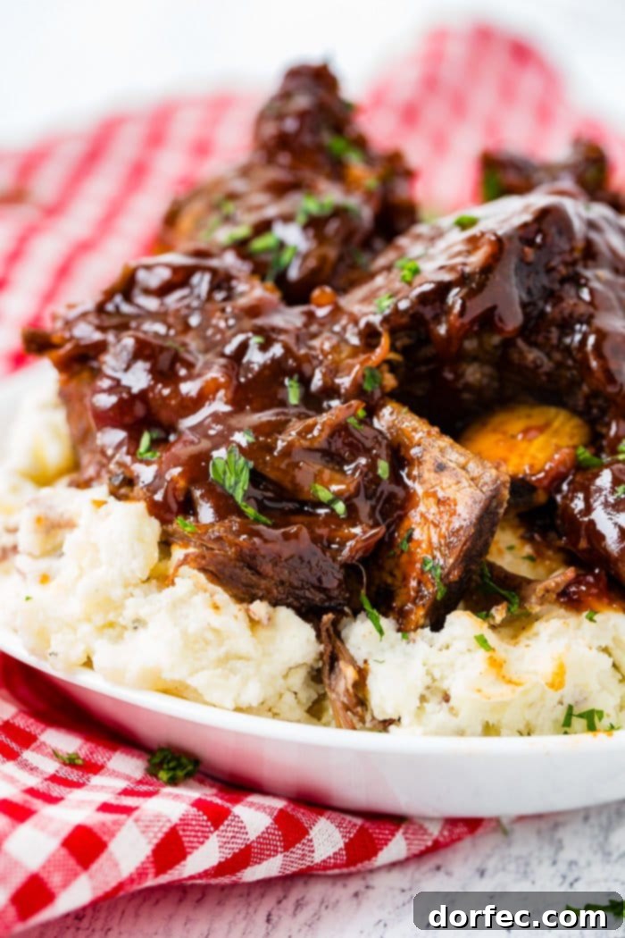 A photo of sticky bbq beef short ribs piled on top of mashed potatoes on a white plate on top of a red and white checkered cloth napkin.
