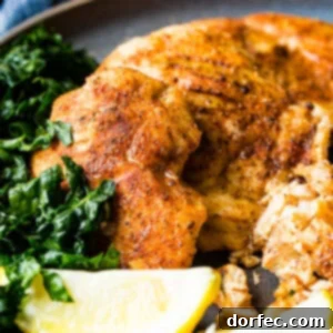 A photo of whole smoked chicken breasts and sliced smoked chicken breasts on a charcoal gray plate with lemon wedges and kale on the side.