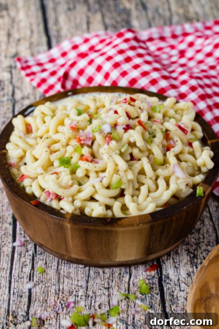 Ultimate Macaroni Pasta Salad Recipe 4 A wooden bowl on a red and white checkered napkin full of creamy cold noodles and a mix of colorful vegetables, perfect for a picnic.