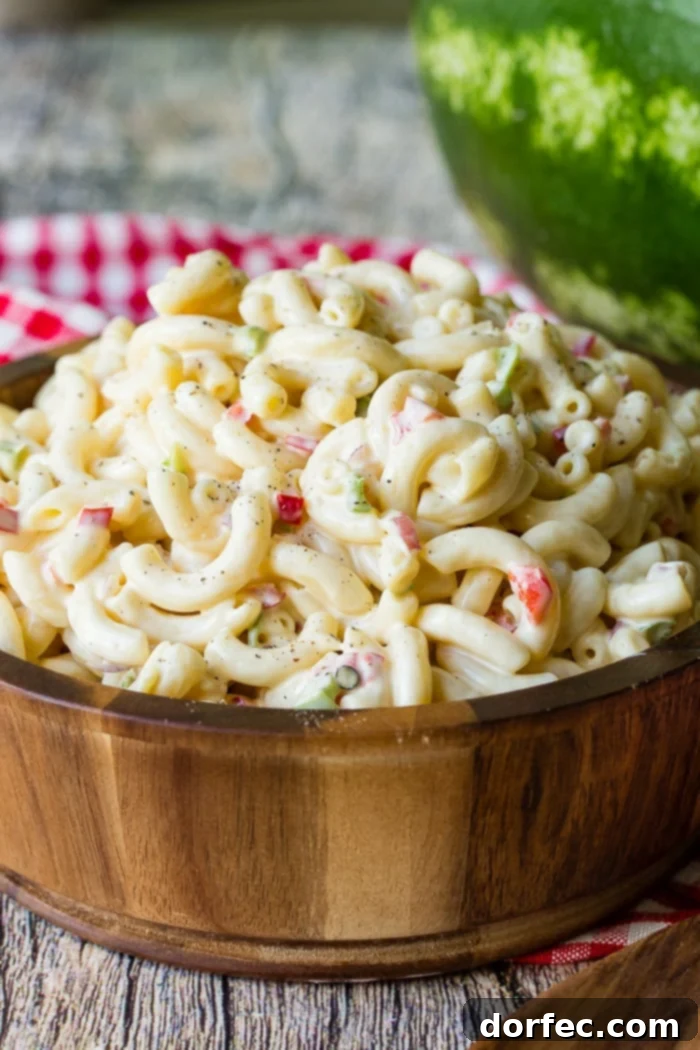 Ultimate Macaroni Pasta Salad Recipe 6 A wooden bowl with a red and white checkered napkin in the background, filled with creamy pasta and crisp vegetables, ideal for summer dining.