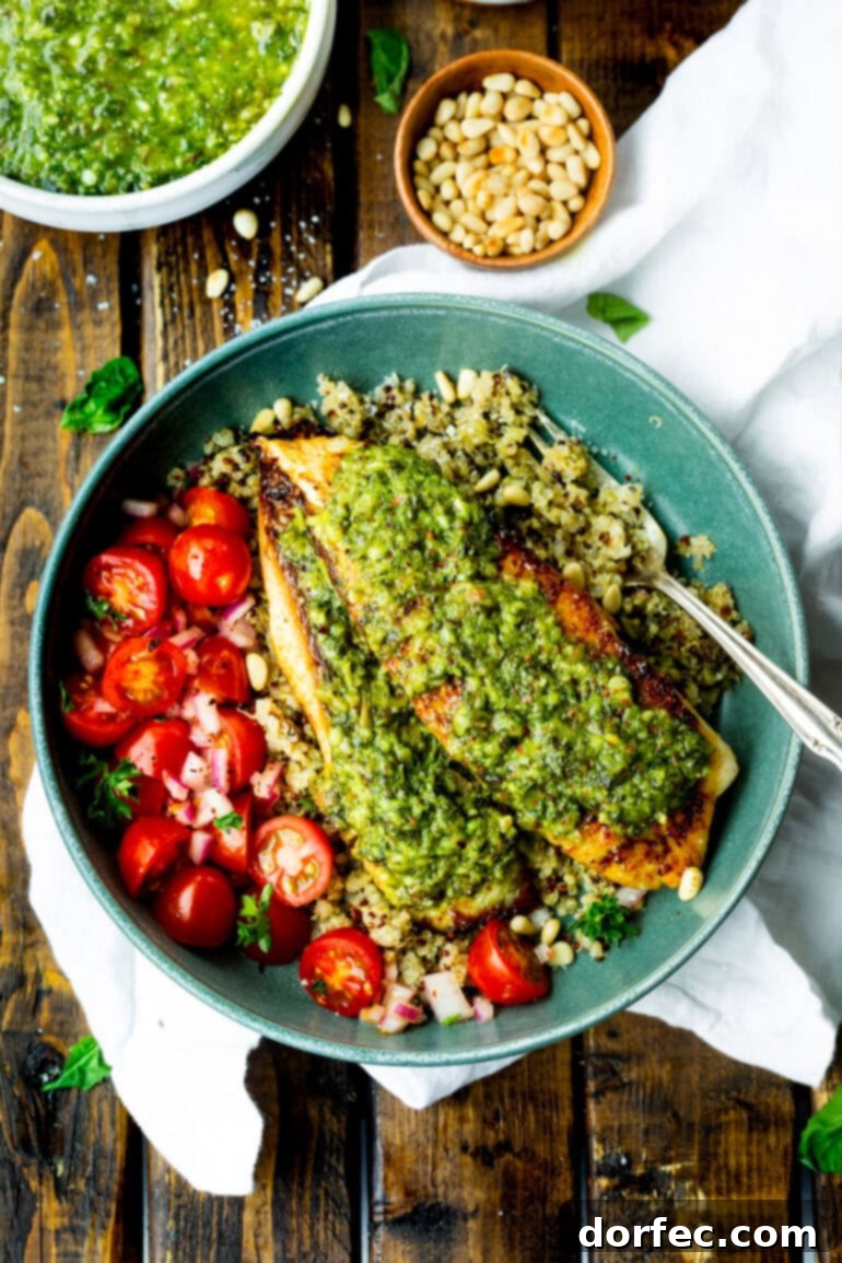 Light and Lean Pesto Tilapia Dinner 4 Two seared tilapia fillets, each beautifully topped with a spoonful of vibrant pesto sauce, served alongside a colorful medley of quinoa and bright cherry tomatoes. A perfect representation of a healthy and quick meal.