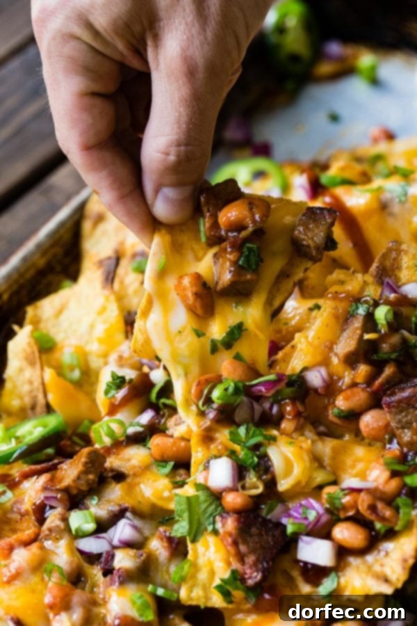 A close-up of a hand pulling a single, perfectly cheesy nacho chip from a pile of BBQ brisket nachos, highlighting the stretchy, melted cheese.