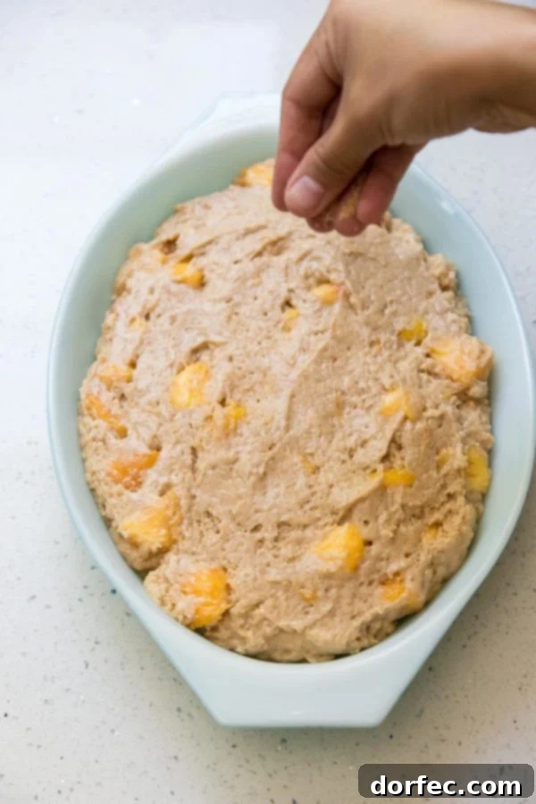 A baking pan filled with unbaked peach dump cake batter, ready to go into the oven. The batter is studded with peach pieces and has a smooth, creamy texture.