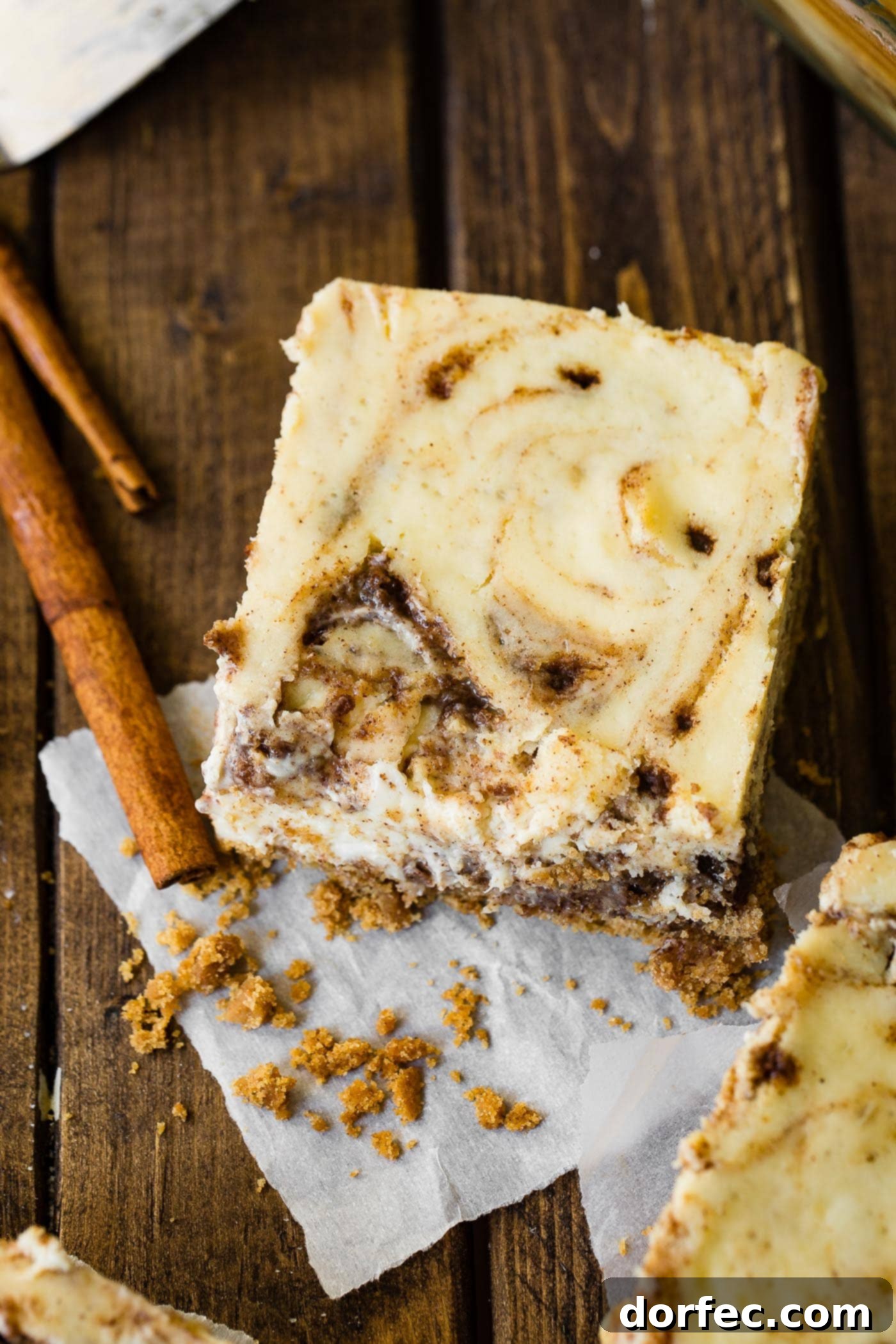 A photo of a cinnamon roll cheesecake bar sitting on a wooden surface with cinnamon sticks sitting next to it, highlighting its rich texture.