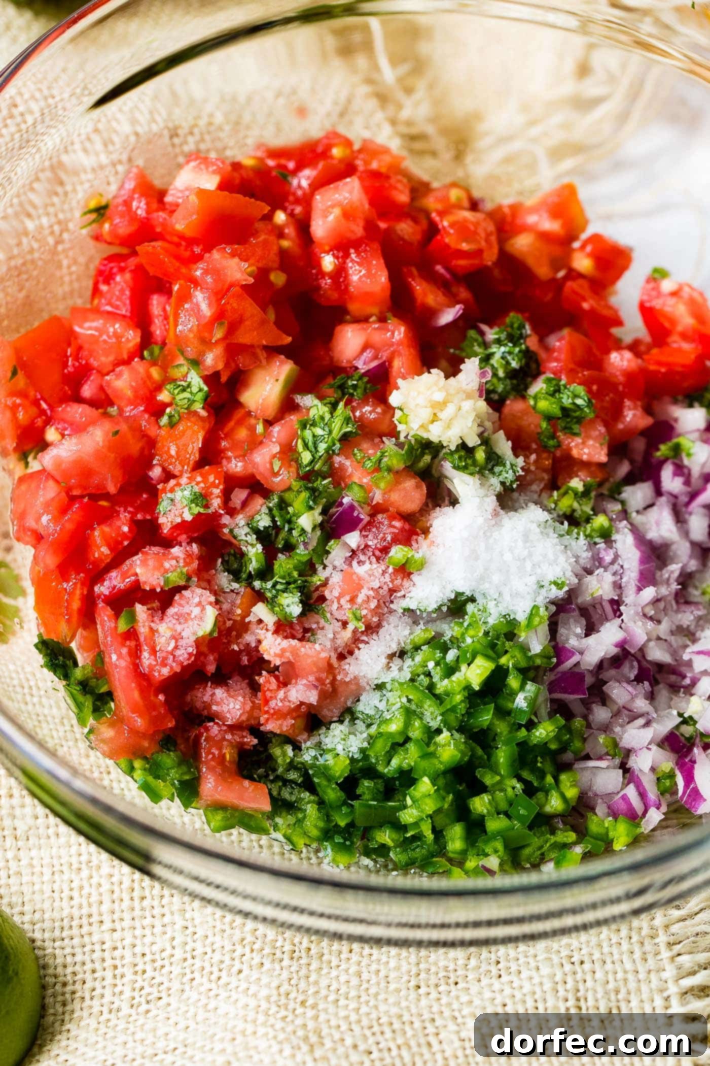 A glass bowl filled with the vibrant, uncooked ingredients for Pico de Gallo – diced tomatoes, red onions, cilantro, and jalapenos – awaiting a final stir.