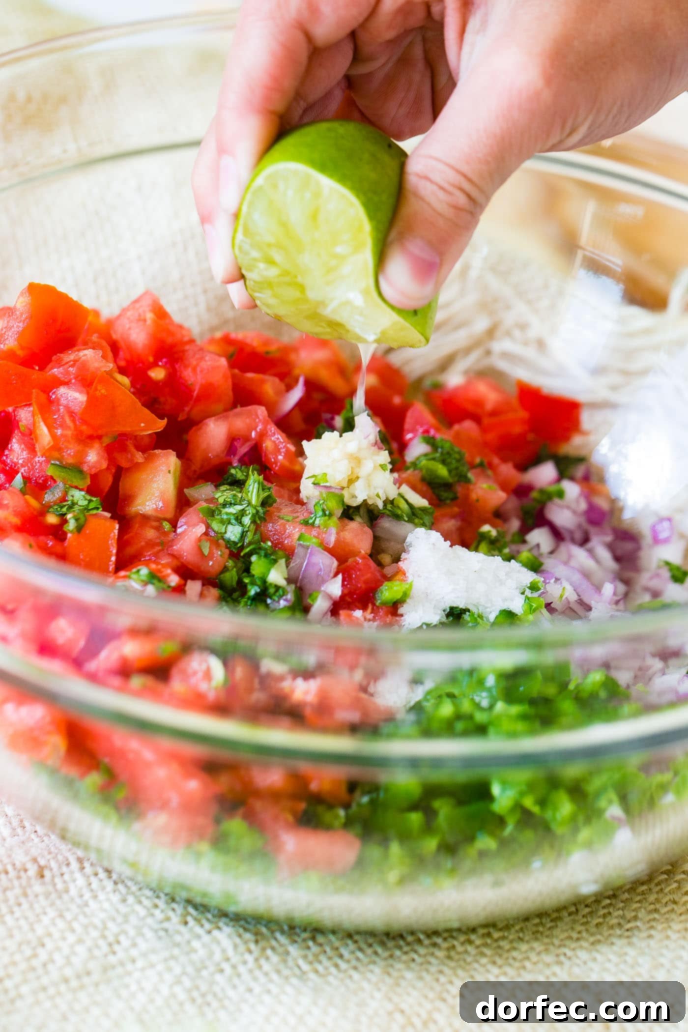 A close-up shot of a glass bowl filled with Pico de Gallo ingredients, with a hand squeezing fresh lime juice over them, highlighting the essential citrus element.
