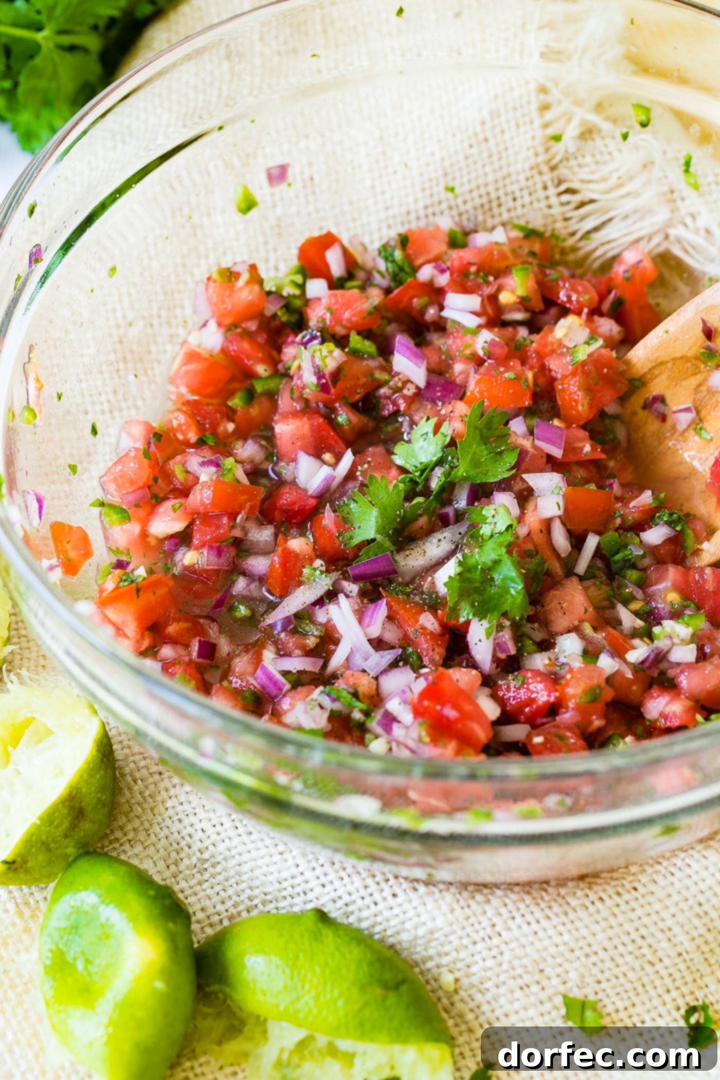 A clear glass bowl of freshly prepared Pico de Gallo, being gently stirred with a wooden spoon to evenly distribute the vibrant, colorful ingredients.