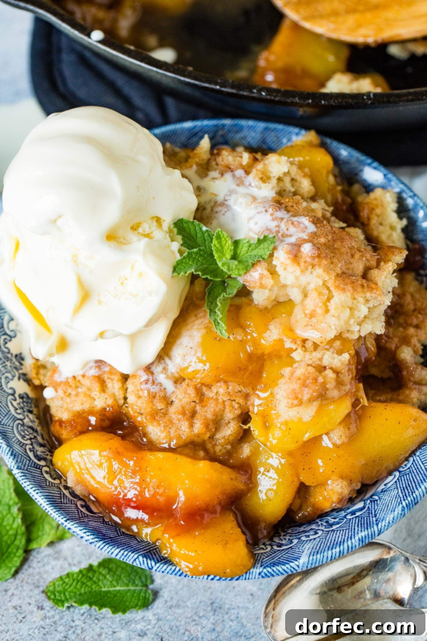 A photo of a bowl of fresh peach cobbler with a scoop of vanilla ice cream on top and a sprig of fresh mint, highlighting the golden biscuit topping and juicy peaches.