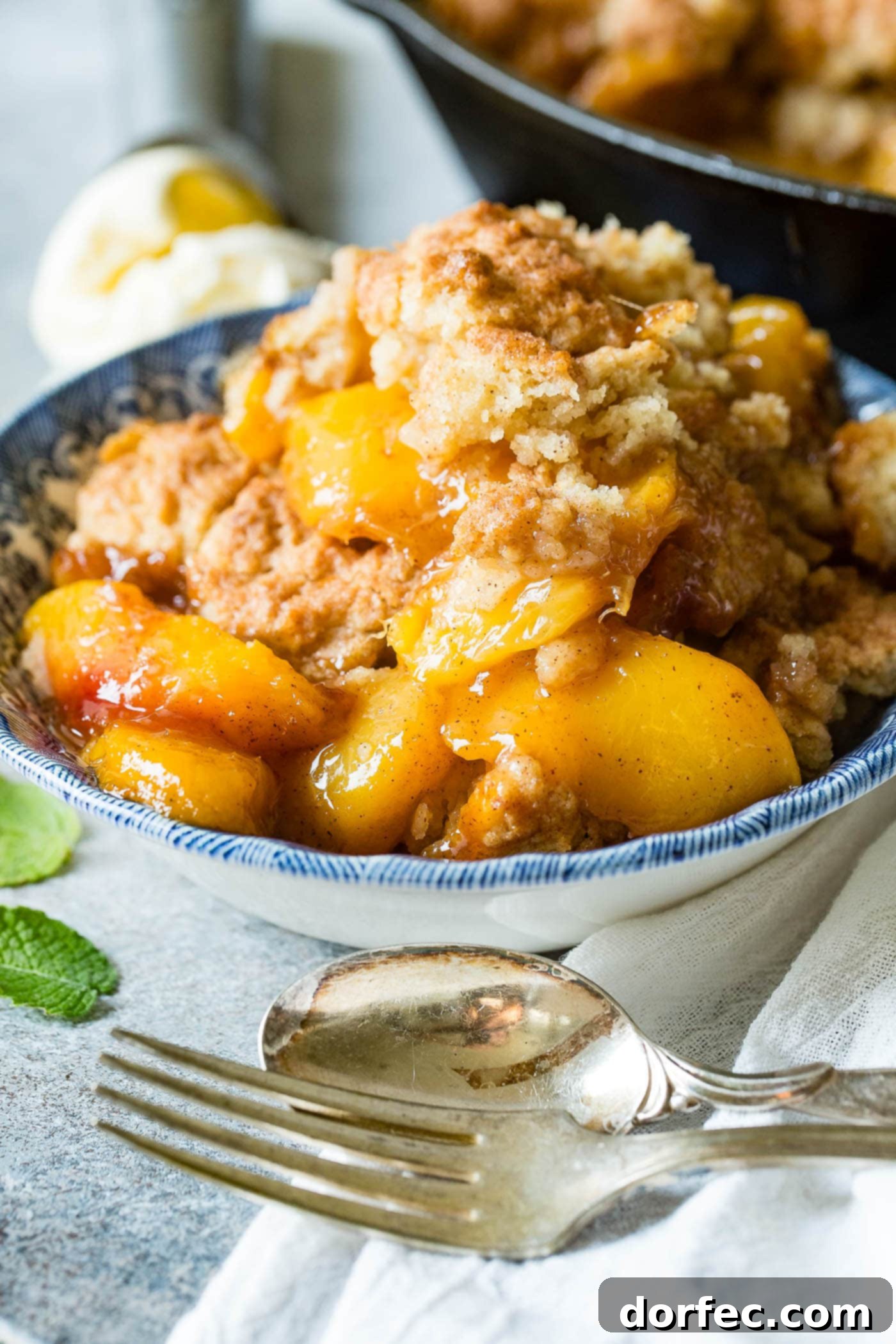 A photo of a bowl of peach cobbler with a biscuit topping, topped with a scoop of vanilla ice cream and a sprig of fresh mint.