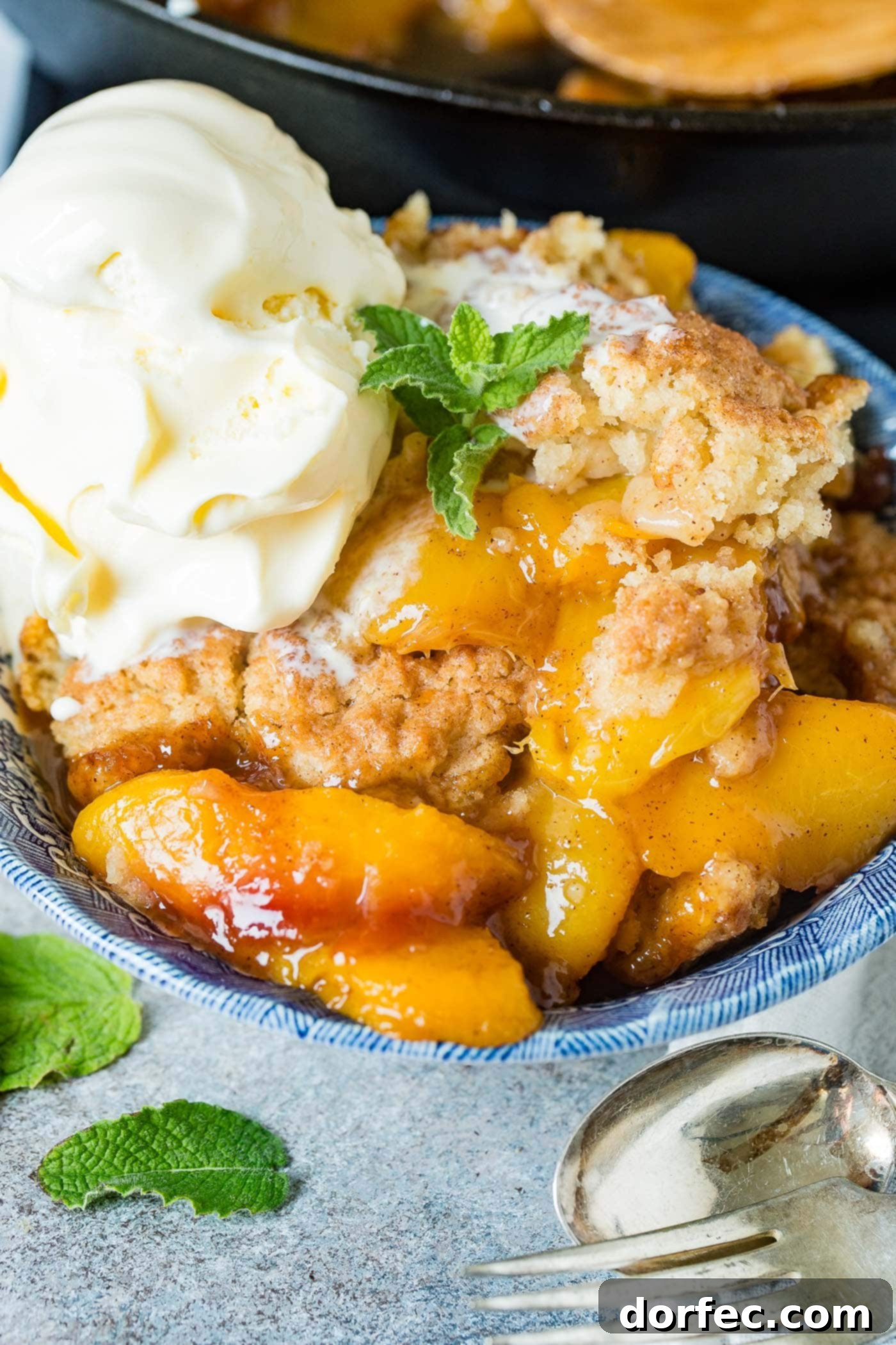 A close-up photo of a bowl of fresh peach cobbler with a generous scoop of vanilla ice cream on top and a sprig of fresh mint, emphasizing the rich textures and appealing presentation.